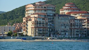 Un hotel de varios pisos con balcones, situado cerca de una playa con gente y barcos.