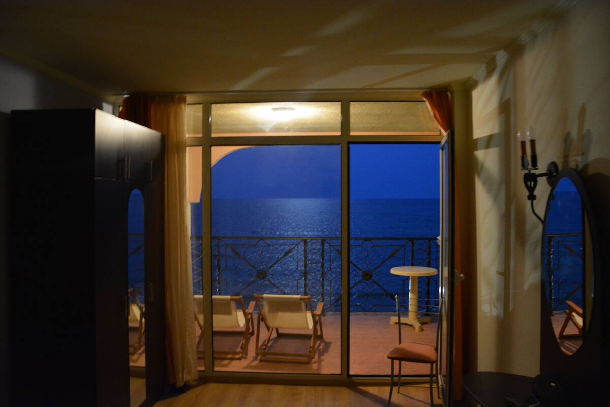 A room with a view of the sea, a balcony with chairs and a table, and a large window.