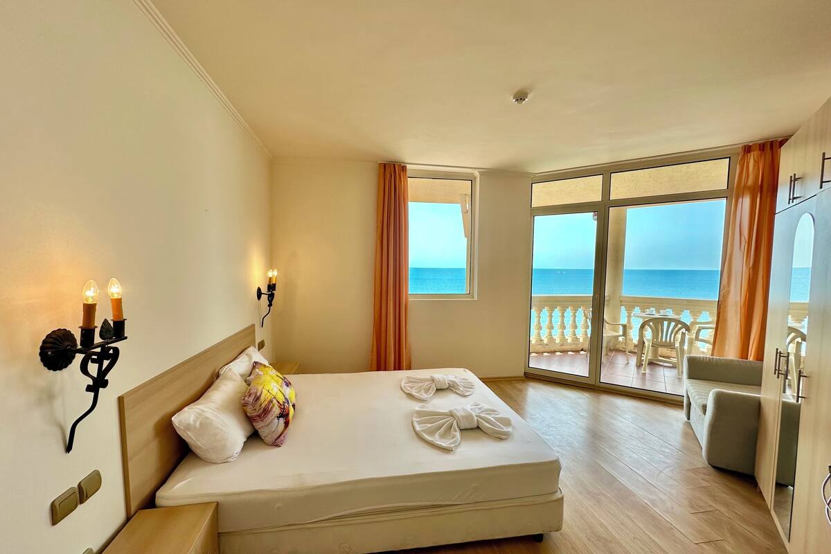 A hotel room with a bed, a nightstand, a wall-mounted light fixture, and a view of the sea through large windows.