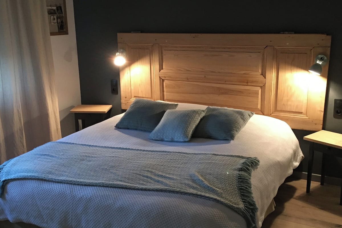 A neatly made bed with a textured blanket and pillows, a wooden headboard with wall-mounted lights, and a small bedside table.