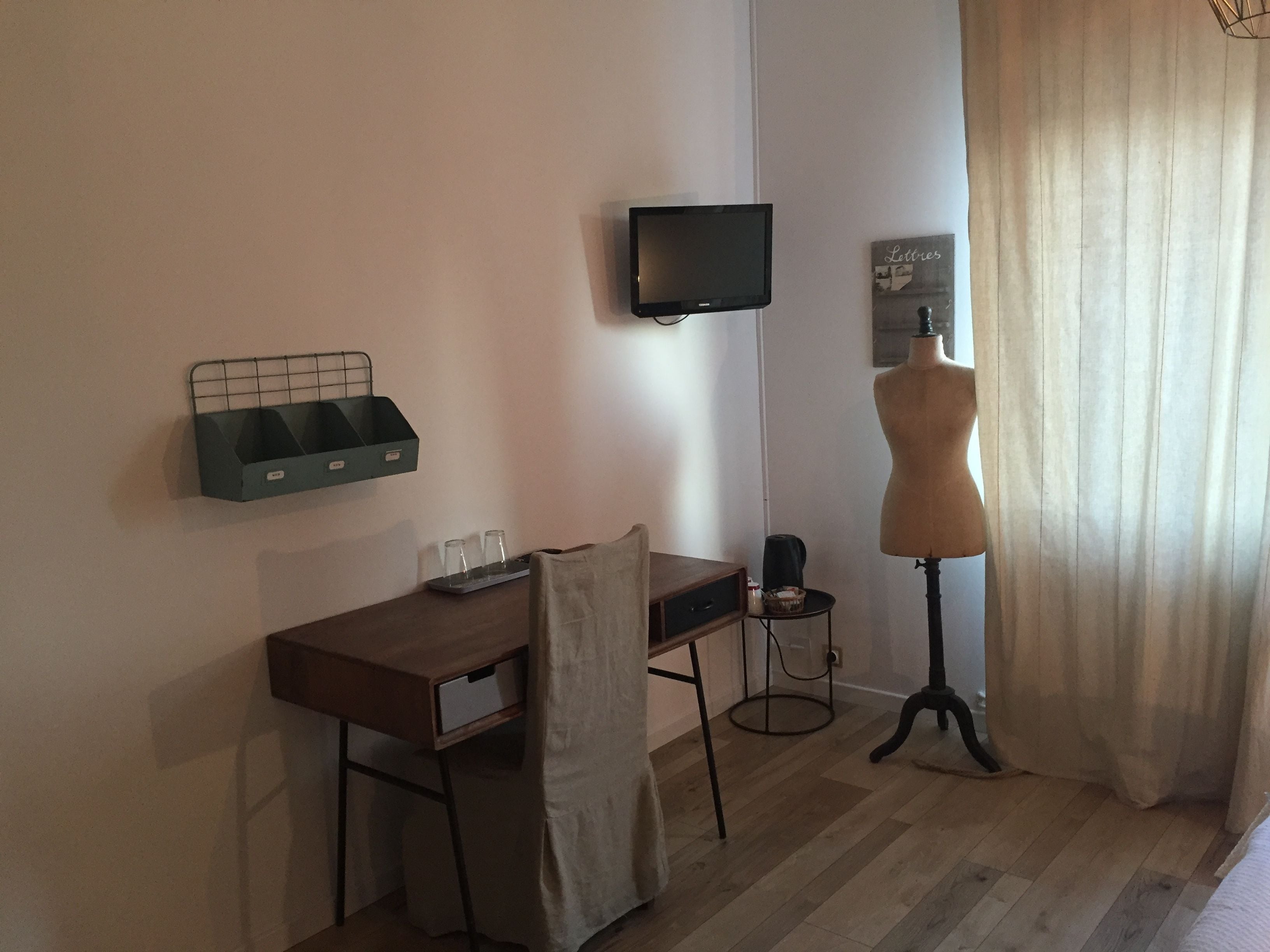 A room with a wooden desk, a chair, a television mounted on the wall, a mannequin, and a window with curtains.