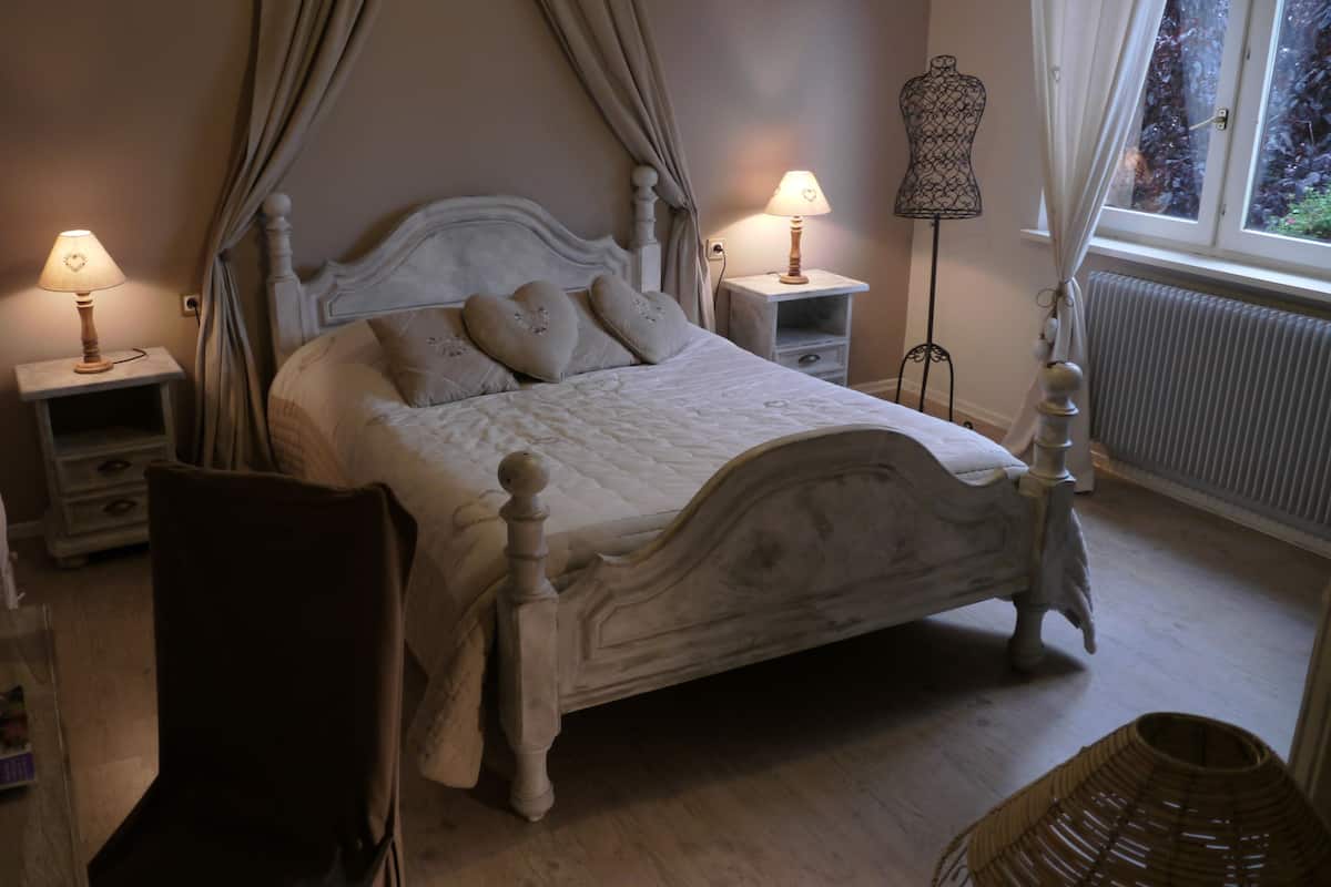 A bedroom with a four-poster bed, two bedside tables with lamps, a chair, a wicker basket, and a radiator.