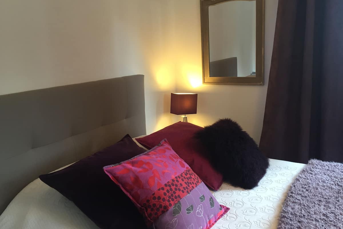 A bed with a grey headboard, a red and purple pillow, a black fluffy pillow, a bedside lamp, and a mirror on the wall.
