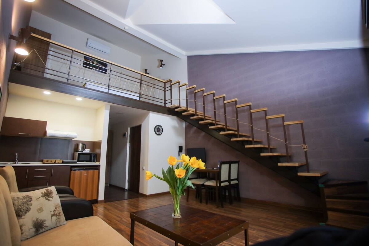 A spacious living area with a wooden staircase, a dining table, and a kitchen in the background.