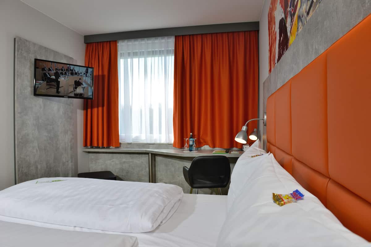 Une chambre d’hôtel avec un lit, un bureau et une chaise. Il y a une fenêtre avec des rideaux orange, un téléviseur fixé au mur et un lit avec du linge de lit blanc.