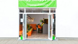 The entrance of Steepy Steepy Hotel with a green sign, glass doors, orange seating, and a reception desk.