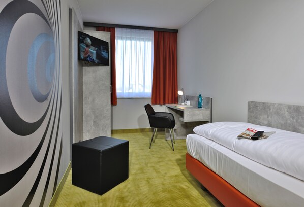Una habitación de hotel con cama, escritorio, silla, televisor y un llamativo diseño abstracto en la pared.