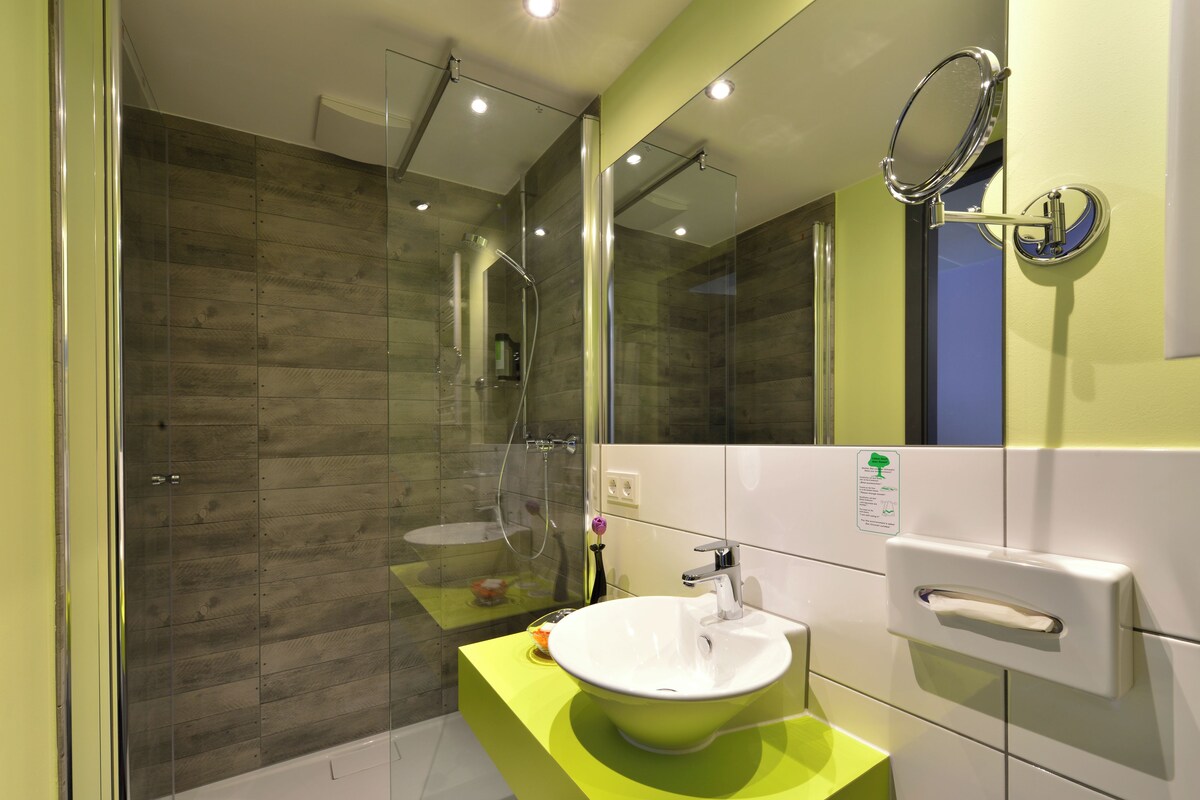 Un baño moderno con ducha envuelta en vidrio, un lavabo blanco con encimera verde y un espejo con marco redondo.