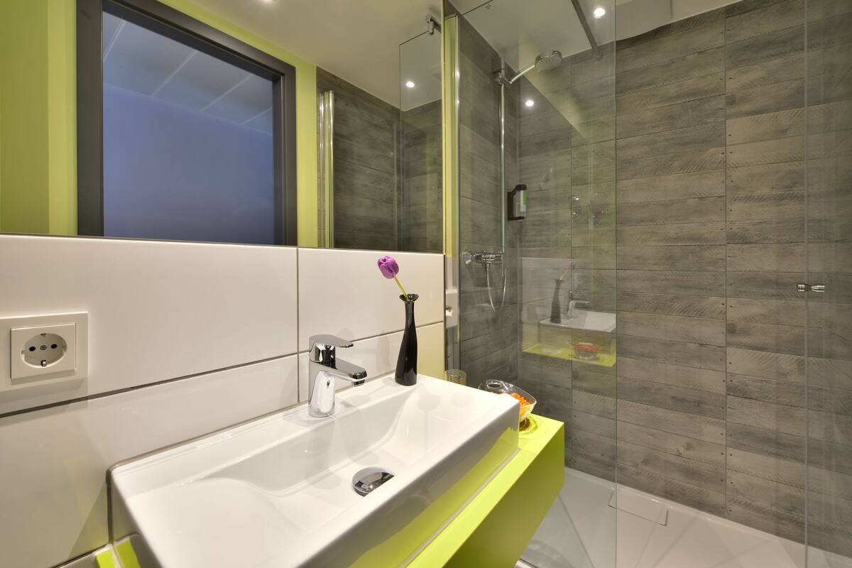 Une salle de bain moderne avec un lavabo blanc, un meuble-lavabo jaune, une cabine de douche en verre et un miroir au-dessus du lavabo.