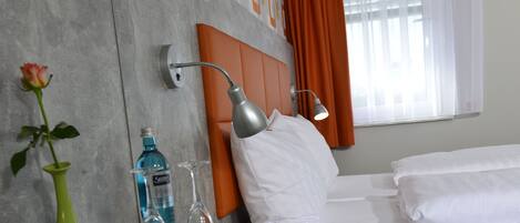 A hotel room with a bed, a nightstand with a lamp, a vase with a rose, a bottle of water, and a glass.