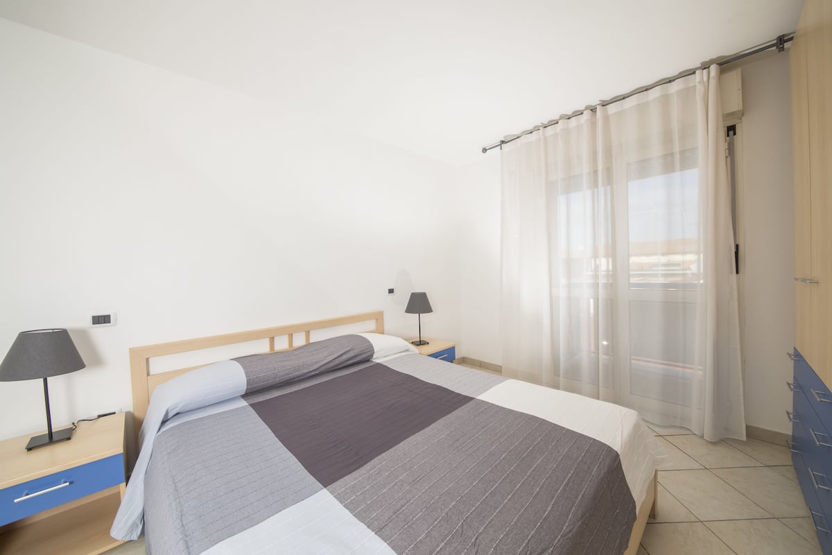 Quarto com cama, mesa de cabeceira, abajur e janela com cortinas.