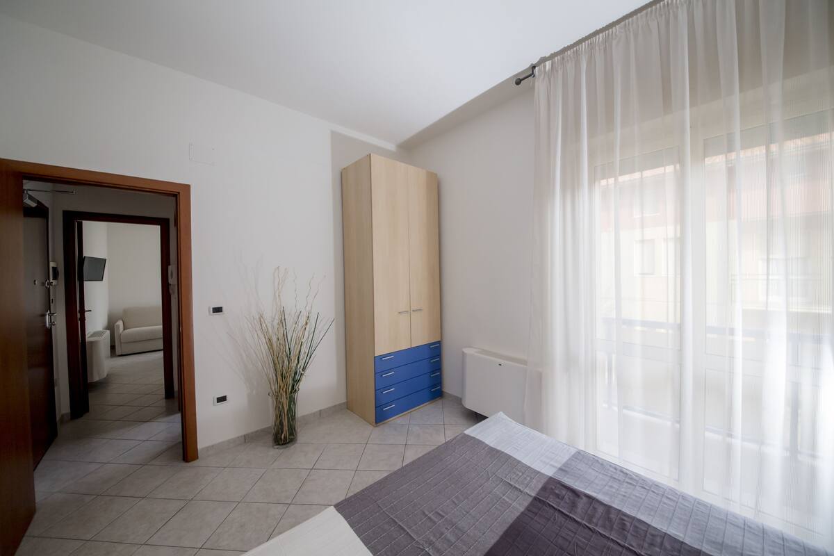 Um quarto com cama, guarda-roupa, radiador e janela com cortinas leves.