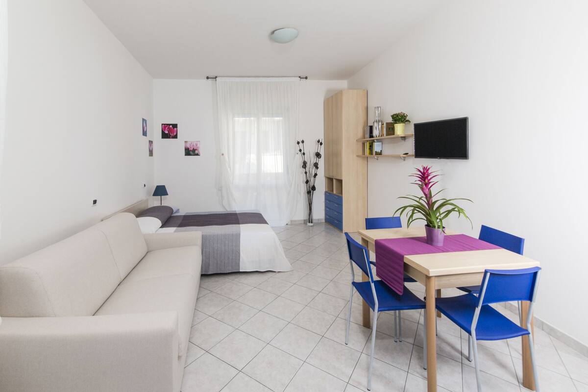 Uma sala de estar moderna e bem iluminada, com um sofá, uma mesa de jantar com cadeiras, uma TV na parede e uma janela com cortinas leves.