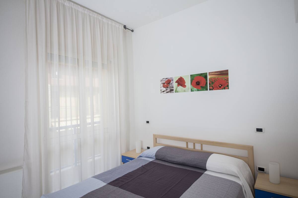 Quarto com cama, mesa de cabeceira, janela com cortinas leves e parede com quatro quadros.
