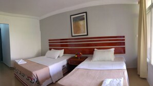 Quarto de hotel com duas camas, cabeceira de madeira, um quadro na parede e janela com cortinas.