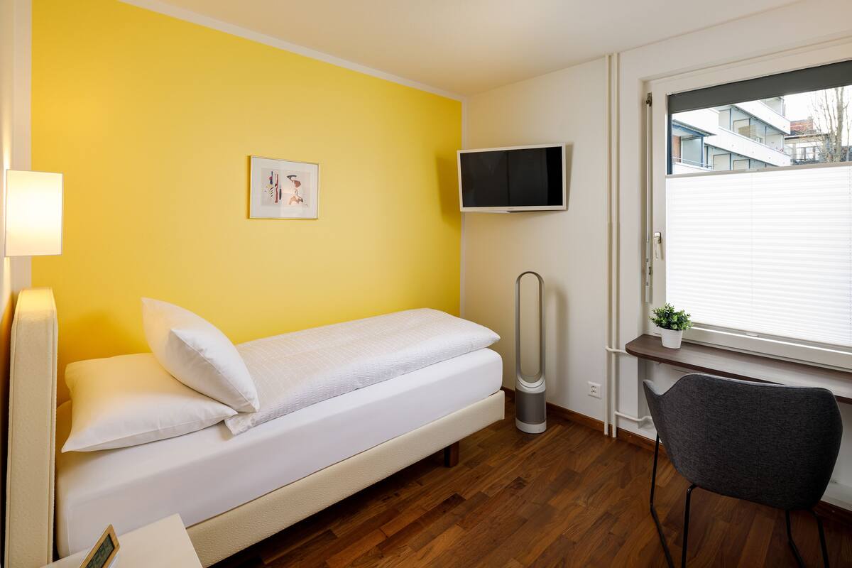 A small room with a bed, a desk, a chair, a TV, and a window with blinds.