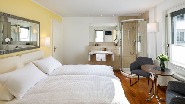 Habitación de hotel con dos camas, baño con ducha, una mesita pequeña y una silla.