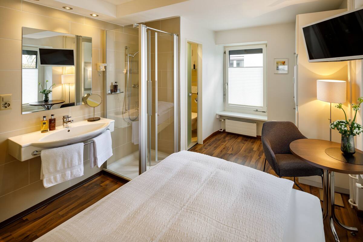 A modern bathroom with a glass shower enclosure, a sink with a mirror, and a bedside table with a lamp.