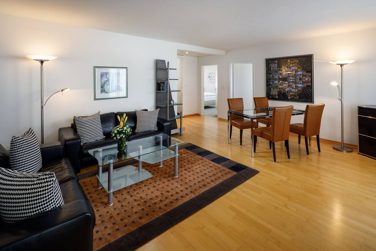 A modern living room with a black leather sofa, a glass coffee table, a dining area with brown chairs, and a large abstract painting on the wall.