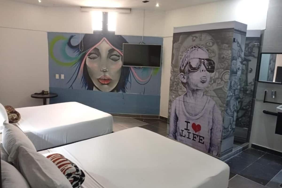 A hotel room with two beds, a TV, a mural on the wall, and a bathroom with a mirror.