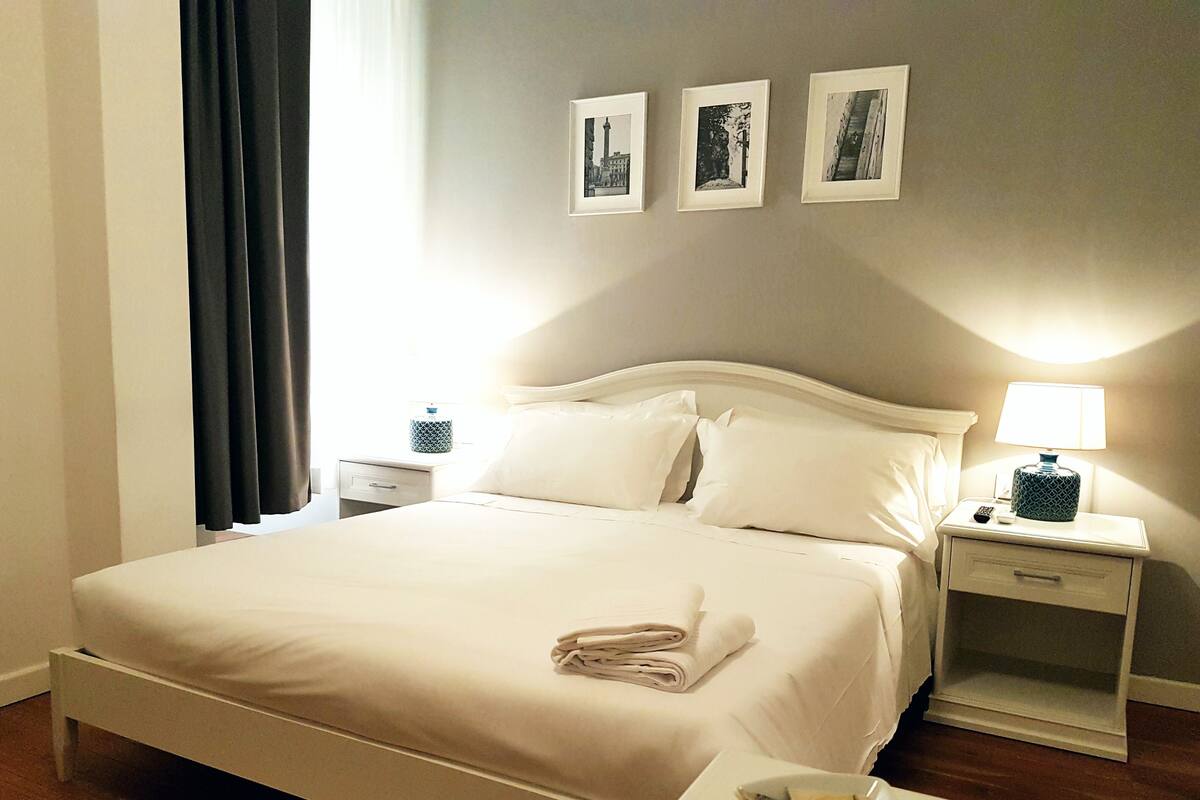 A hotel room with a neatly made bed, a bedside table with a lamp, a small bedside table with a decorative item, and three framed pictures on the wall.