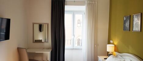 Une chambre d’hôtel comprenant un lit, un bureau, une chaise, une télévision, une fenêtre avec des rideaux et une table de chevet avec une lampe.