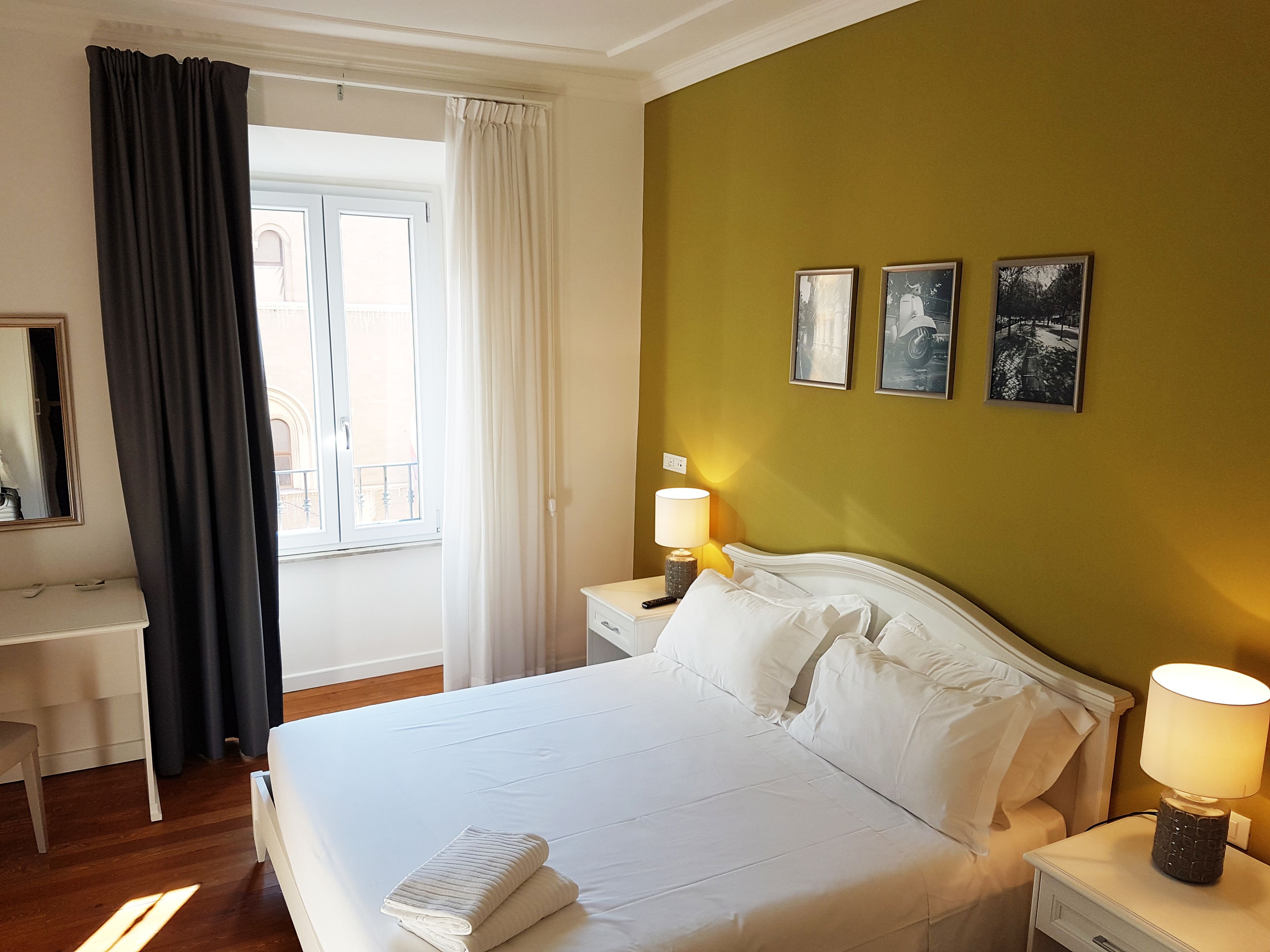 Une chambre d’hôtel comprenant un lit, un bureau, une chaise, une fenêtre avec des rideaux et trois tableaux encadrés accrochés au mur.