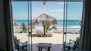 Vistes a la platja des d'una habitació amb portes corredisses de vidre, incloent un para-sol de palla, dues cadires i una taula petita.