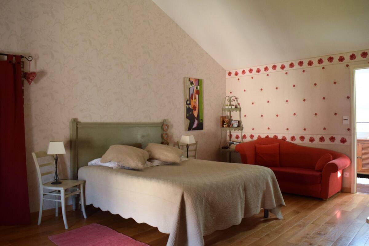Un dormitorio con cama, sofá rojo, silla, mesita de noche, lámpara y un papel tapiz floral en la pared.
