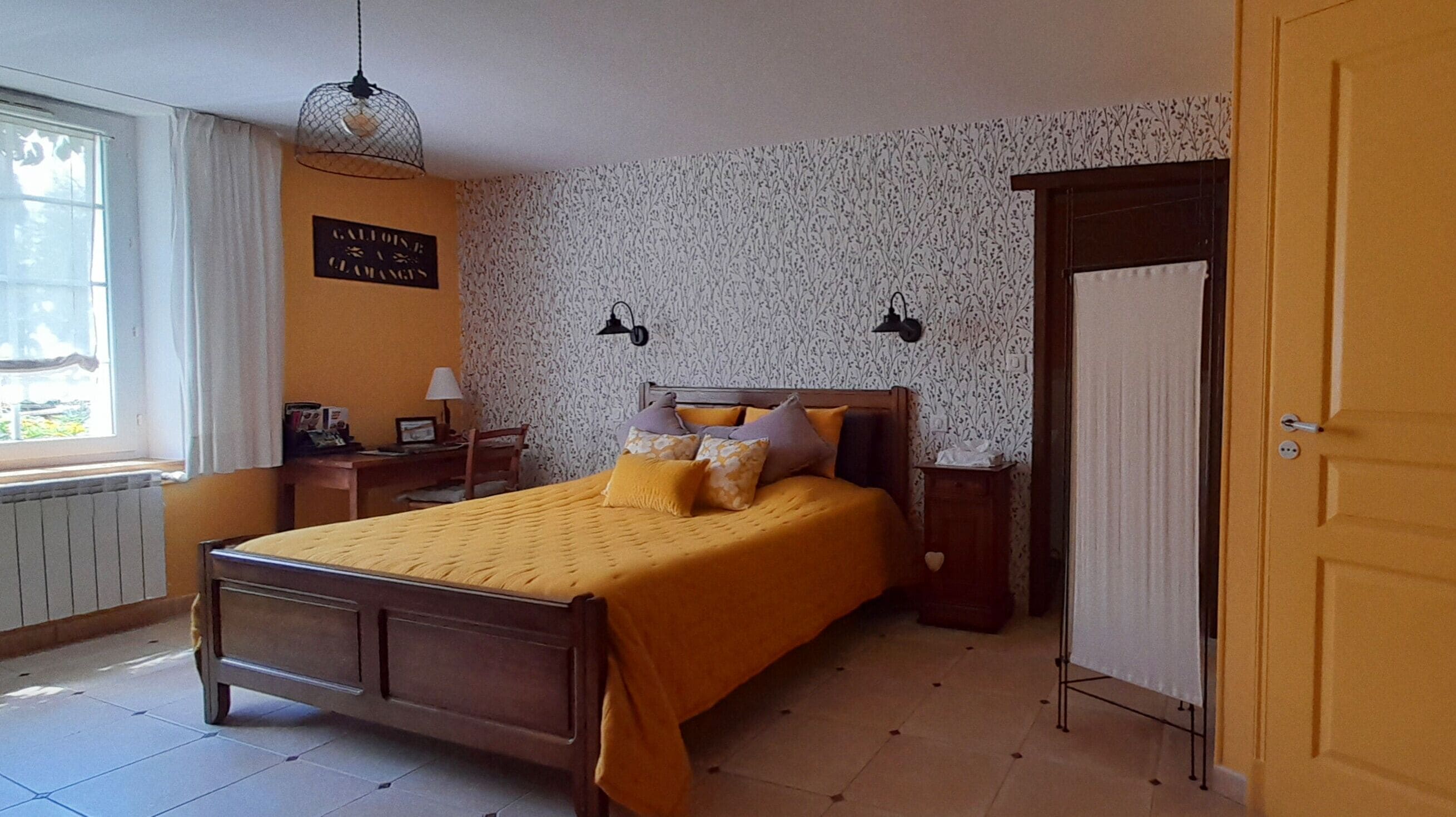 Een slaapkamer met een bed, nachtkastje, lamp, raam met gordijnen, radiator, deur en een muur met bloemenprint.