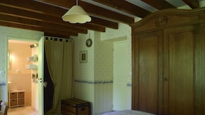 Un dormitorio con un armario de madera, una cama con ropa de cama blanca, un baúl de madera y un baño visible a través de un puerta abierta.