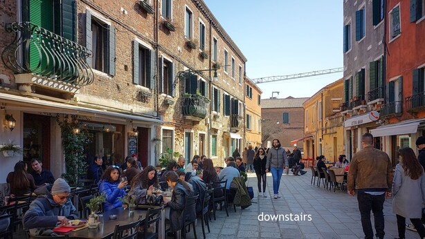 A bustling outdoor café area with tables and chairs, surrounded by buildings with balconies and windows. - Cross Canal Set (Venice)