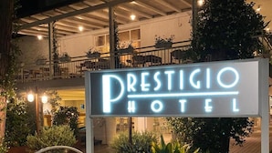 Une enseigne pour l’hôtel Prestigio, avec une entrée bien éclairée et un espace jardin agrémenté de fleurs et de plantes.
