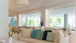 Un salon moderne avec un canapé beige, de nombreux coussins, une table basse blanche et une plante en pot. De grandes fenêtres laissent entrer la lumière naturelle et offrent une vue sur l’extérieur.
