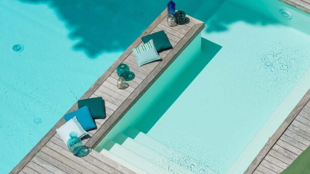 Une terrasse en bois avec une piscine, comprenant un espace détente avec des coussins et un livre.