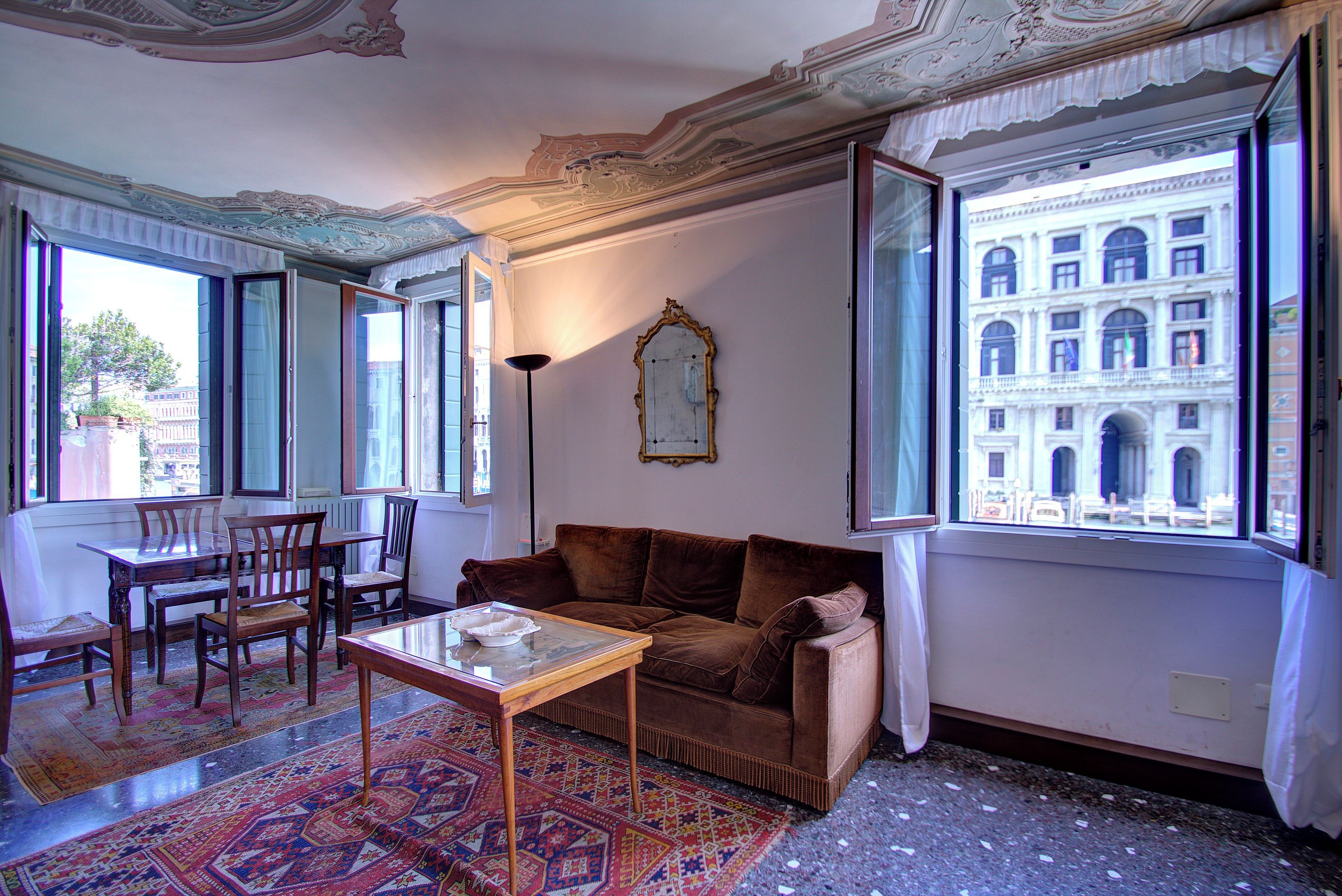 A room with a brown sofa, a wooden table, and a patterned rug. There is a large window with a view of a building.