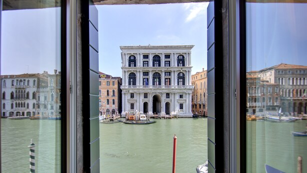 A view of a historic building with a large arched entrance, a canal with boats, and a red pole in the water. - GRITTI Grand Canal (Venice)