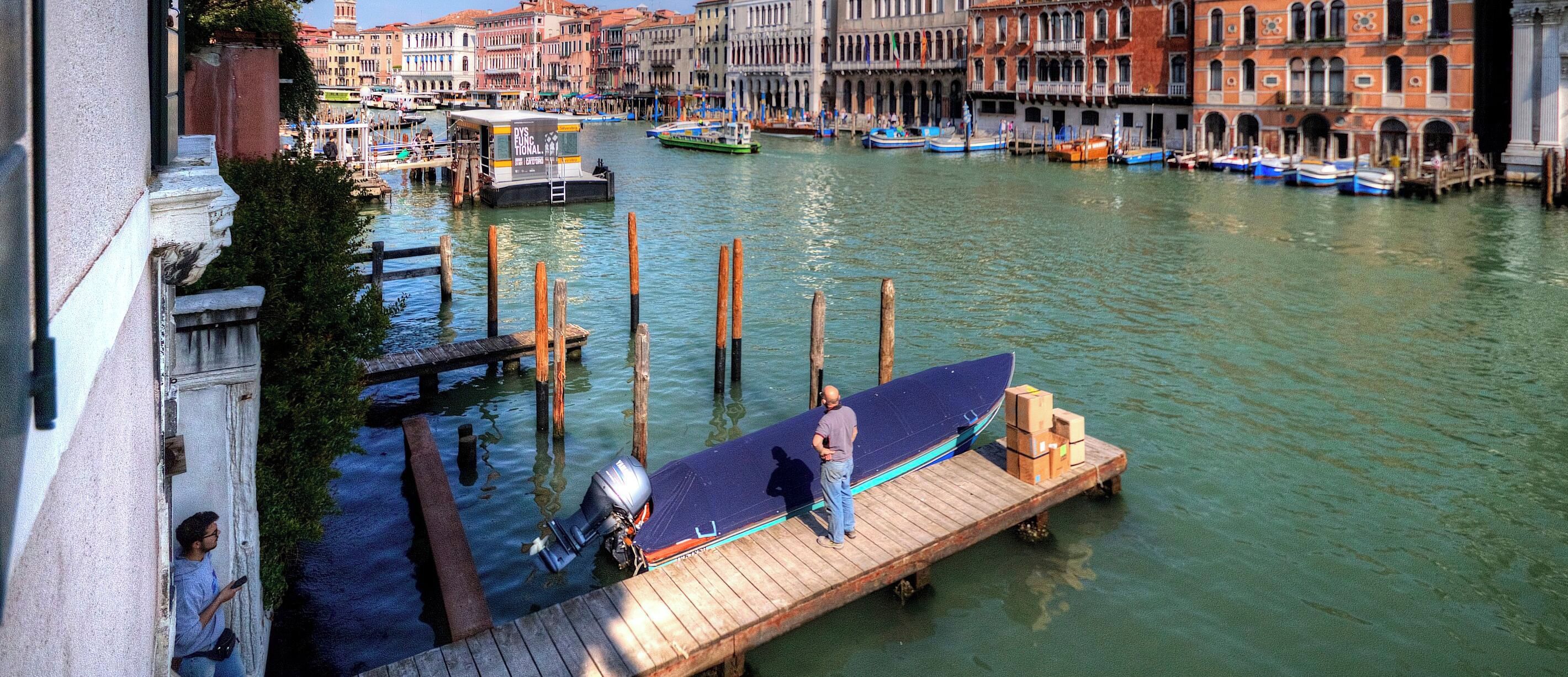 A canal with boats docked, a wooden pier, and buildings in the background.
