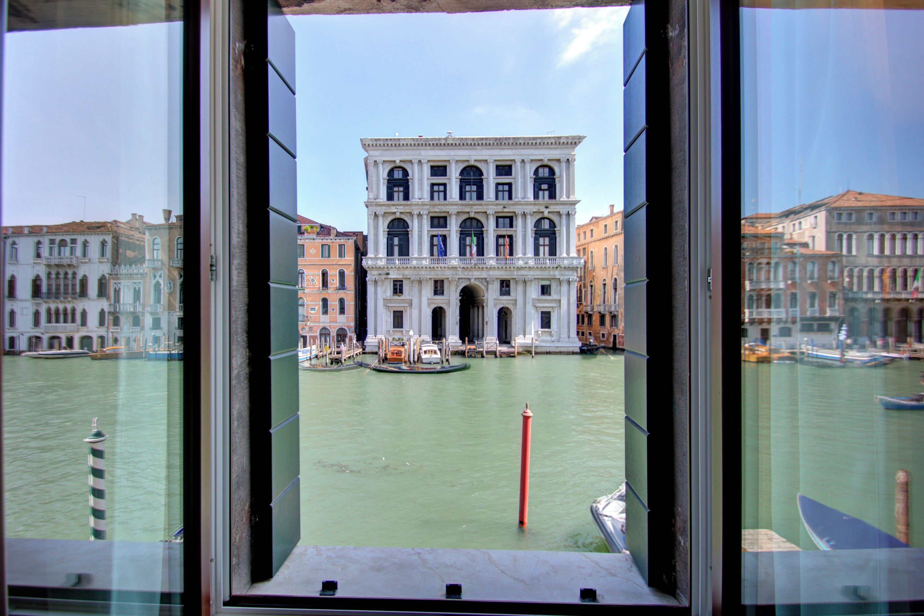 Blick auf ein historisches Gebäude mit großem Bogenportal, einen Kanal mit Booten und einen roten Pfosten im Wasser.