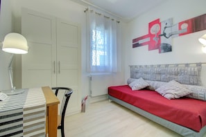 A bedroom with a bed, a desk with a lamp, a window with curtains, and a wall with abstract art.