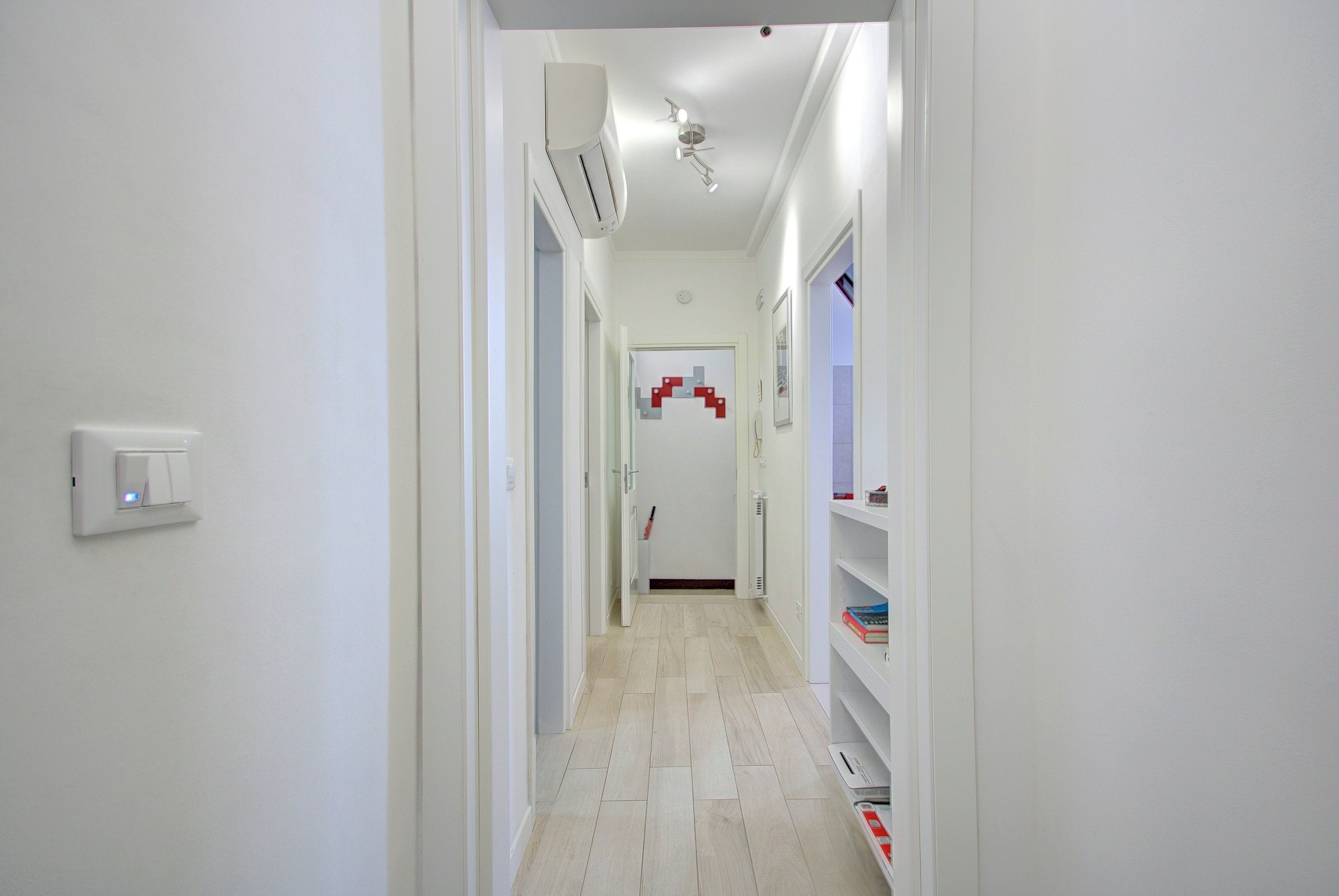 A narrow hallway with a white wall, a light switch, and a white shelf with books.