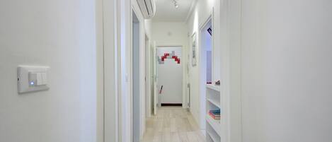 A narrow hallway with a white wall, a light switch, and a white shelf with books.