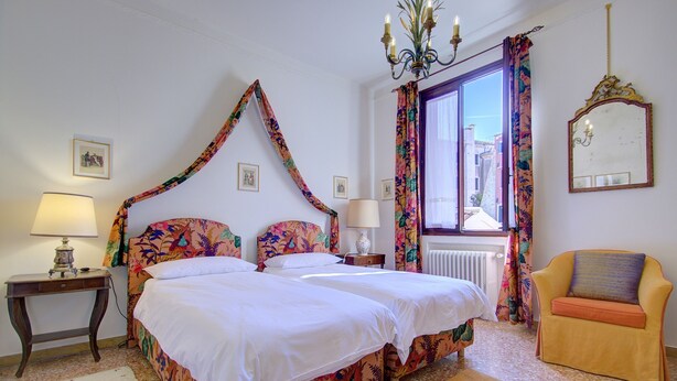 A double bed with floral bedding, a bedside table with a lamp, a chair, a window with curtains, and a mirror on the wall.