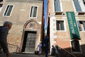 A narrow street with a historic building featuring a large wooden door and a green banner with white text. - BAROCCO-Veneziano (Venice)