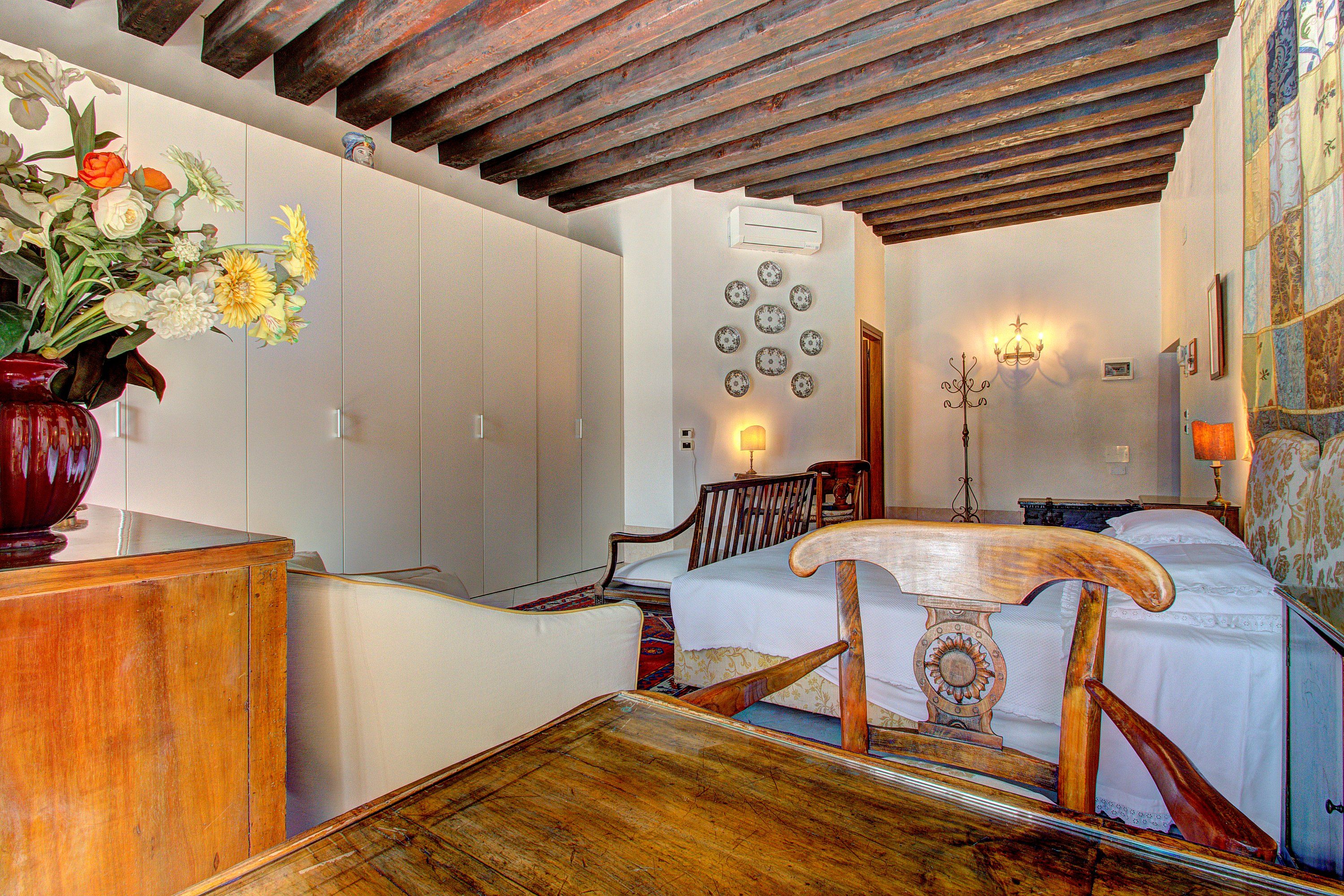 A bedroom with a wooden bed frame, a wooden dresser, a vase with flowers, and a wall-mounted air conditioner.