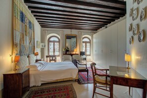 A bedroom with a large bed, a desk, and a chair. There are two windows with arched tops and a mirror above the bed. - TRIBUNO Grand Canal (Venice)
