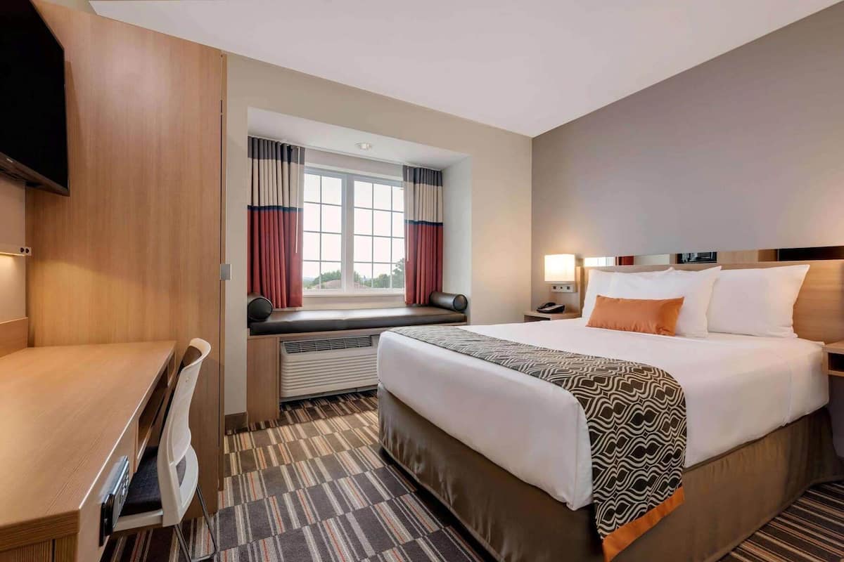 Habitación de hotel con cama, escritorio, silla, ventana con cortinas y televisor.