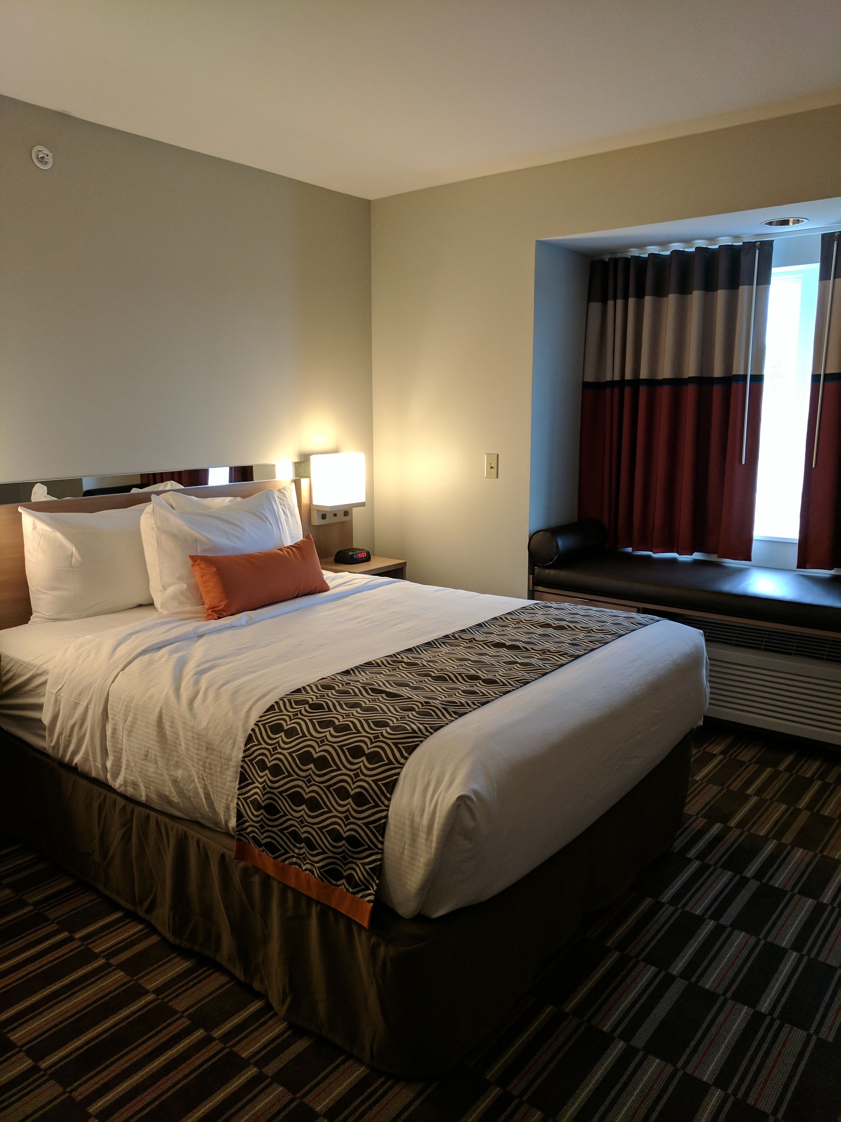 Habitación de hotel con una cama grande, una mesita de noche con lámpara, una ventana con cortinas y suelo alfombrado.