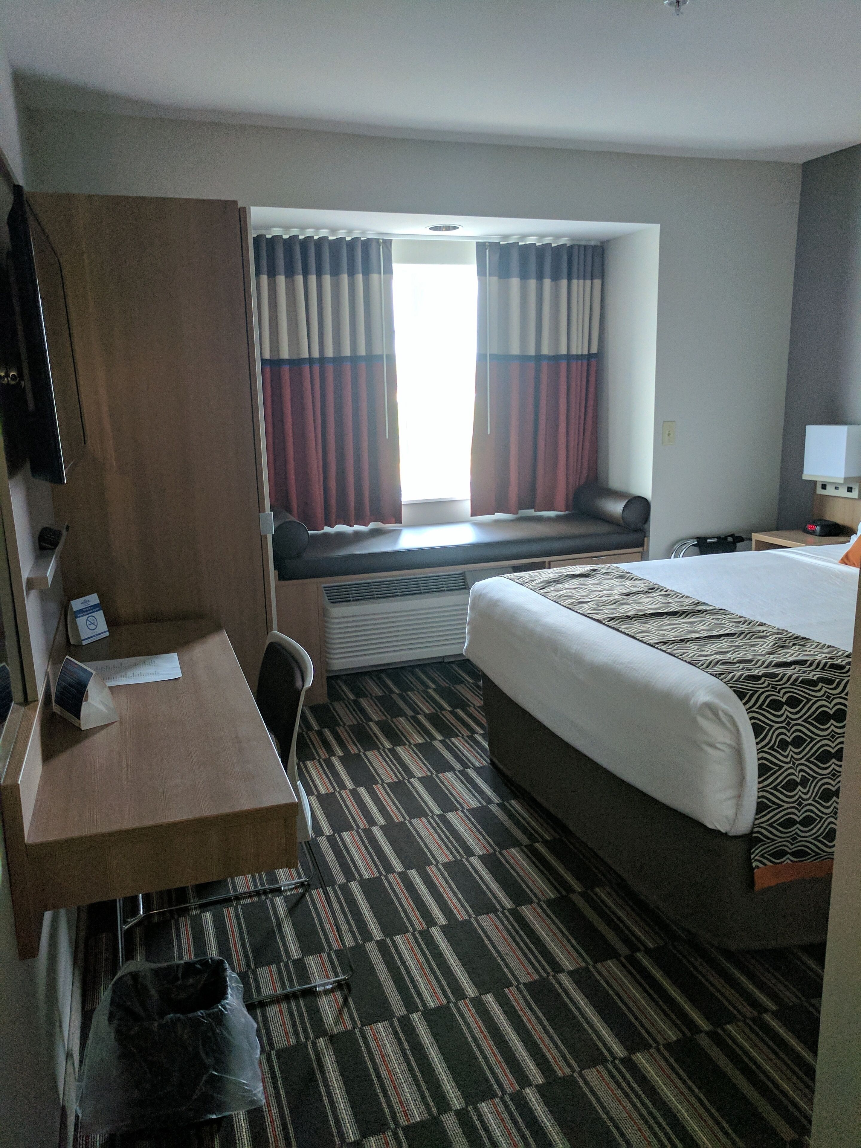 A hotel room with a bed, a desk, a chair, a window with curtains, a heater, and a TV.