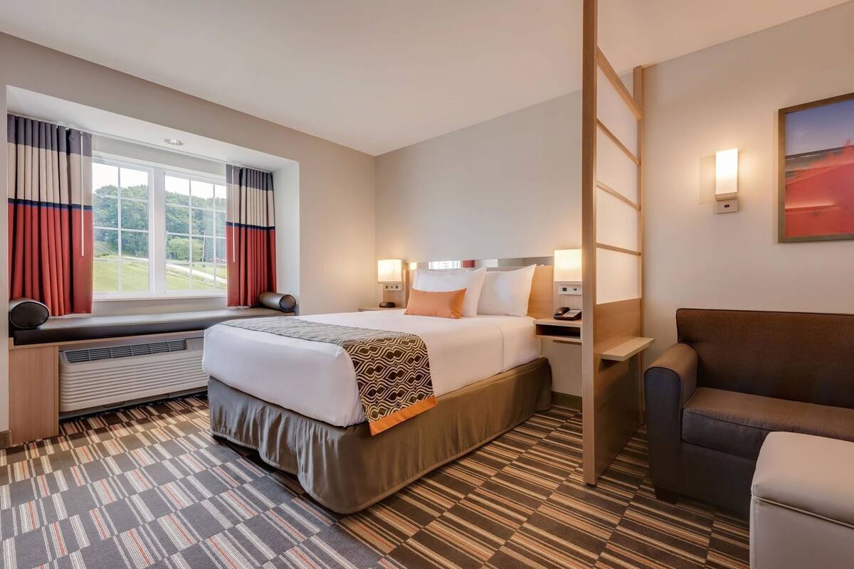 Una habitación de hotel con cama, escritorio, silla, calefacción, ventana con cortinas y un cuadro en la pared.